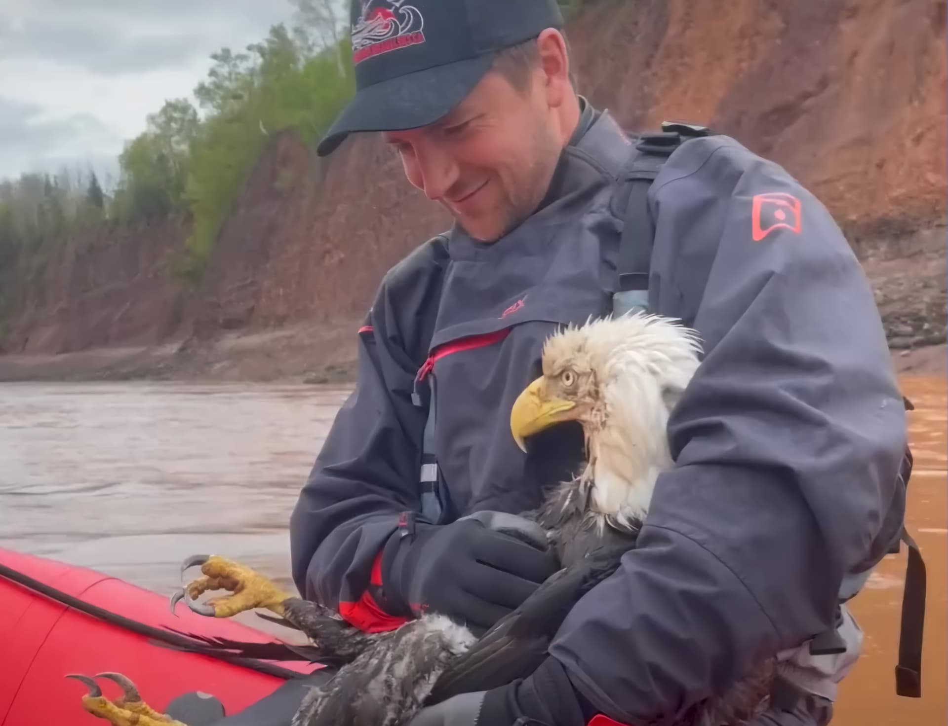 Emmett Blois holding a soaked bald eagle against his chest in a red raft on a muddy tidal river in Nova Scotia